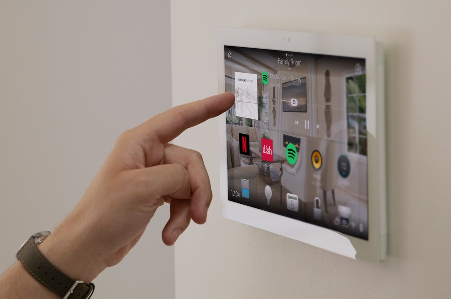A person using a wall-mounted smart home control panel with a Control4 touch screen interface displaying various subsystems. 