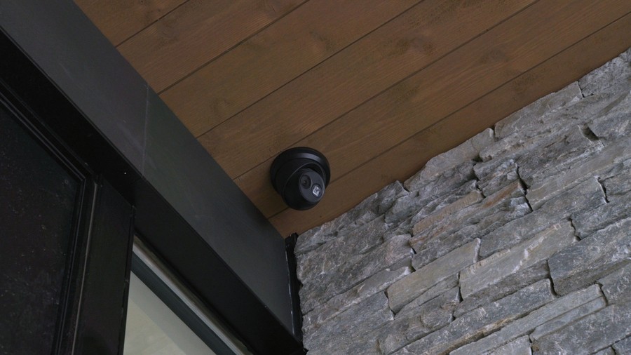 An outdoor security camera mounted on a wooden ceiling next to a stone wall.