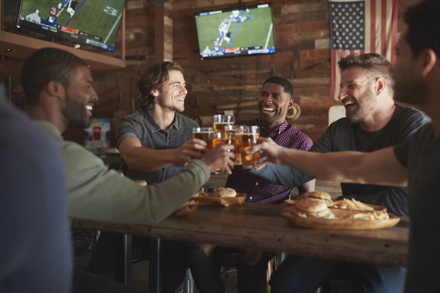 A group of men clink beverage glasses while watching football in a bar.